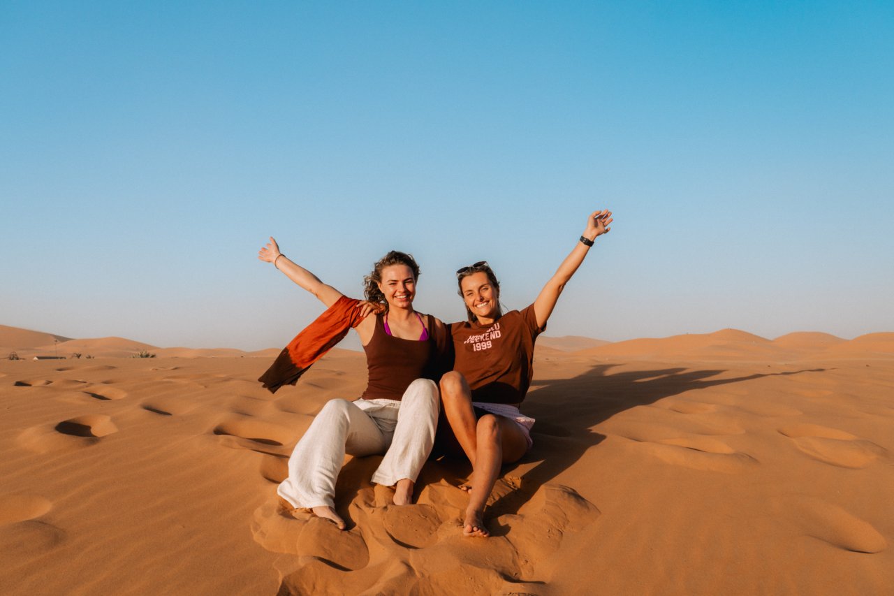 Two young female travellers pose in front of the sand dunes as the sun sets on the Moroccan sand.